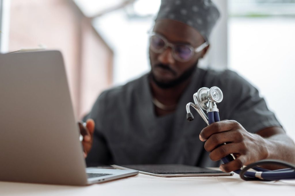 a-man-in-gray-scrub-suit-holding-stethoscope-while-typing-on-his-laptop-5327915 Focused African American doctor in scrubs working with laptop and stethoscope.