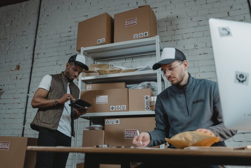 man-in-black-button-up-shirt-standing-beside-man-in-gray-polo-shirt-6169650 Two warehouse employees organizing and packing parcels for delivery in a modern indoor setting.